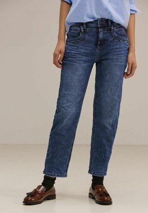 Personne portant un jean bleu, une chemise bleu clair, des chaussettes noires et des mocassins marron à pampilles se tient sur un fond uni clair.