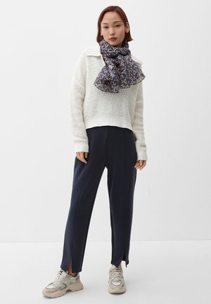 Weißer, flauschiger Pullover mit geripptem Saum, marineblaue Hose mit seitlichen Schlitzen und gemustertes Tuch in Blau, Rot und Gelb. Beigefarbene Sneakers runden das Outfit ab.