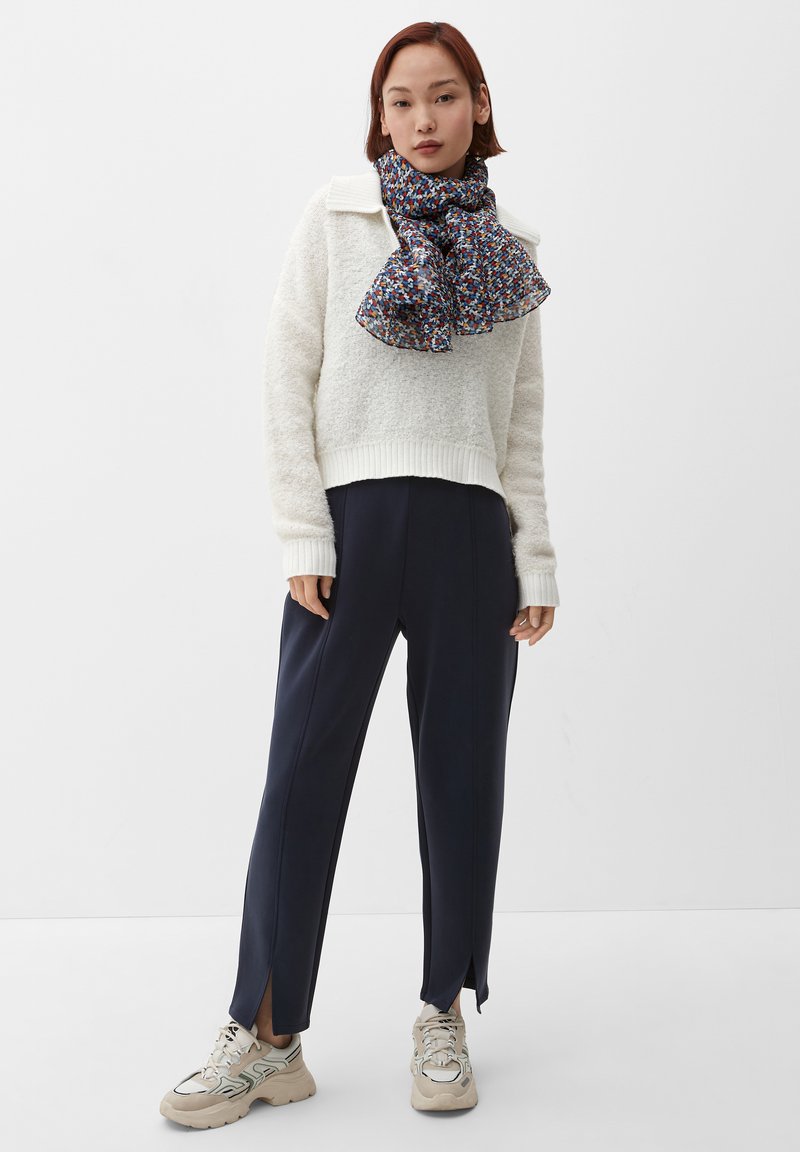 Pull blanc en peluche avec ourlet côtelé, pantalon bleu marine avec fentes sur les côtés et écharpe à motifs bleu, rouge et jaune. Des baskets beige complètent l'ensemble.
