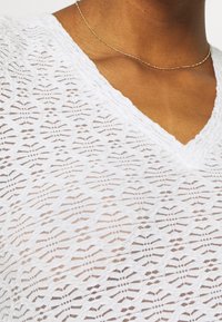 Bílý v-neck top vyrobený z lehké krajky s geometrickým vzorem, který obsahuje texturované linie a malé kulaté akcenty.