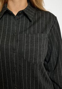 Camicia nera con bottoni e righe verticali bianche, con colletto a punta e taschino sul petto. Realizzata in tessuto texturizzato.