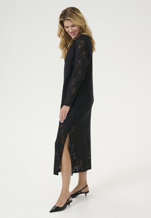 Long black lace dress with long sleeves, front slit, and a fitted silhouette, paired with black slingback heels for an elegant look.