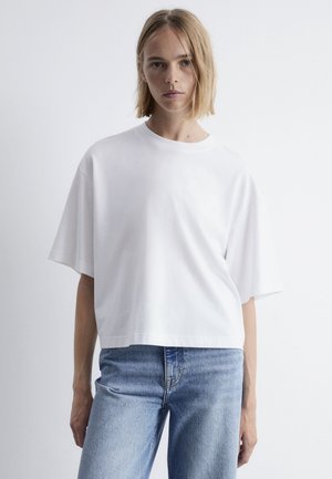 Weißes, kurz geschnittenes T-Shirt mit kurzen Ärmeln, Rundhalsausschnitt und glatter Baumwollstruktur, kombiniert mit hellblauen Jeans. Schlichtes Design.