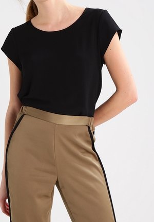 Femme porte un haut noir à manches courtes, rentré dans un pantalon taille haute couleur sable avec des bandes latérales noires sur un fond blanc uni.