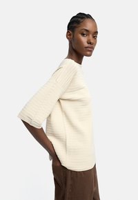 Jasnokremowy sweter z prążkowanego materiału z szerokimi rękawami, o pudełkowym kroju i miękkiej fakturze, w połączeniu z ciemnobrązowymi spodniami.