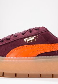 Burgundy mockask sneaker med orange rand, guldlogo och genomskinlig gummiyttersula. Har texturerad yta och vadderad tunga.