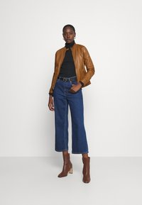 Brown leather jacket with a collar, black turtleneck, blue wide-leg jeans, and brown heeled ankle boots. Model stands against a white background.
