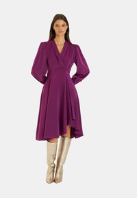 Lila Wickelkleid mit langen Puffärmeln und asymmetrischem Saum, kombiniert mit metallischen kniehohen Stiefeln. Weiche Textur und fließendes Design.