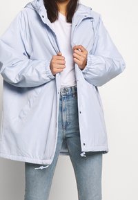 Leggera giacca con cappuccio di colore blu chiaro, con polsini elasticizzati, chiusura a snap e cordini regolabili all'orlo. Indossata sopra una camicia bianca e jeans.