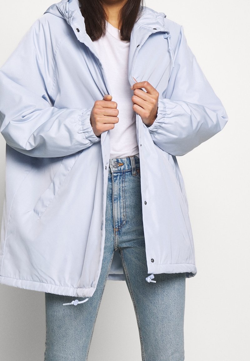 Leggera giacca con cappuccio di colore blu chiaro, con polsini elasticizzati, chiusura a snap e cordini regolabili all'orlo. Indossata sopra una camicia bianca e jeans.