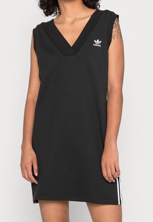 Kobieta ubrana w czarną, bezrękawnikową sukienkę Adidas z dekoltem w serek i białym logo na piersi, widoczne tatuaże na lewym ramieniu.