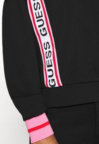 Černý rukáv mikiny s růžovým a červeným lemem, s vertikálním bílým pruhem a černým textem "GUESS" po straně.