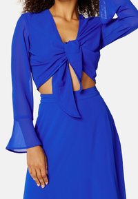 Femme portant un crop top bleu à manches longues avec un nœud à l'avant et une jupe taille haute assortie, montrant son ventre et levant le bras.