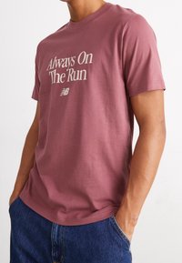 Man in een rozenkleurig T-shirt met "Always On The Run" en een New Balance-logo, gecombineerd met blauwe jeans, met zijn handen in zijn zakken.