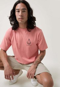 Camiseta de algodón rosa con logo de ancla negro y texto. Modelo vistiendo pantalones cortos beige y zapatillas blancas, agachado contra un fondo neutro.