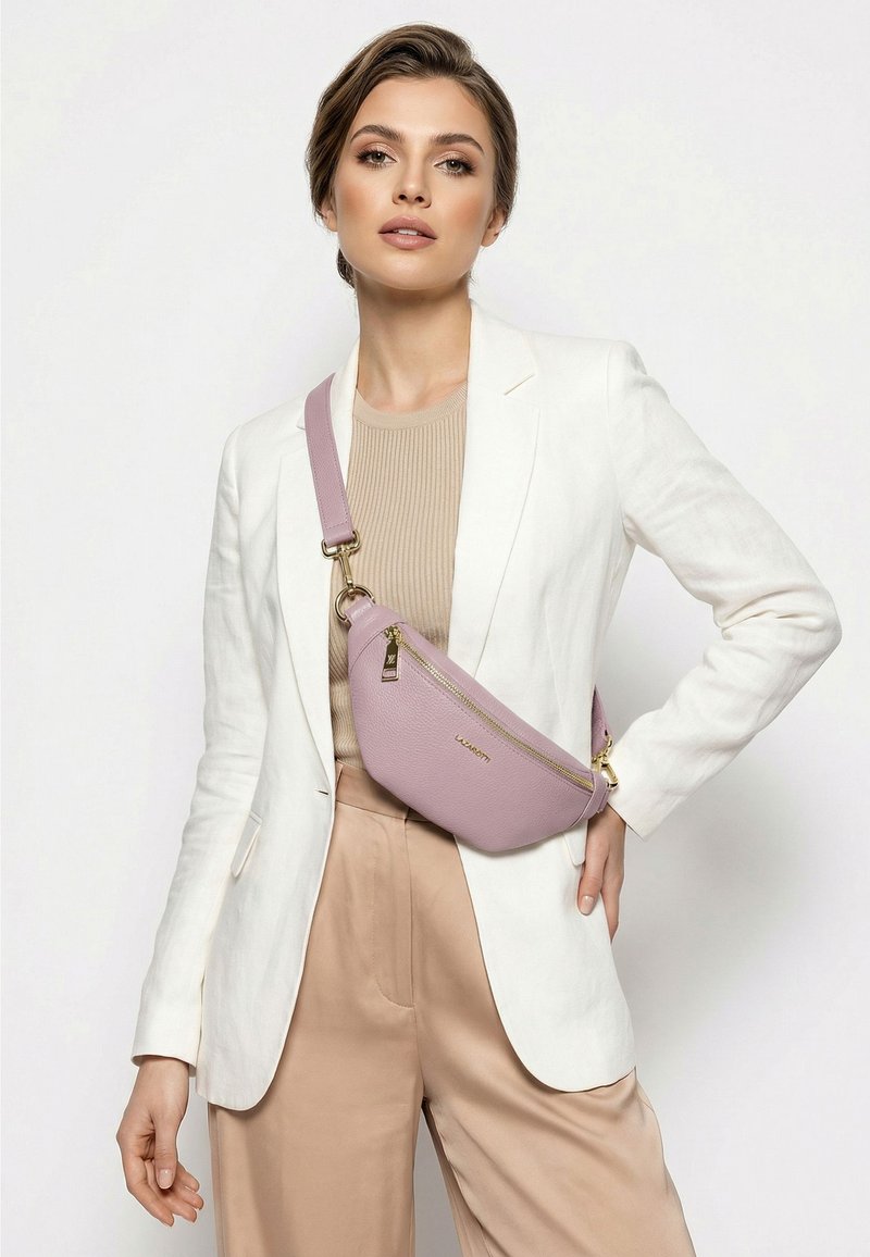 Mujer con blazer blanco y pantalones beige, llevando un bolso bandolera color morado claro con cremallera y herrajes dorados.
