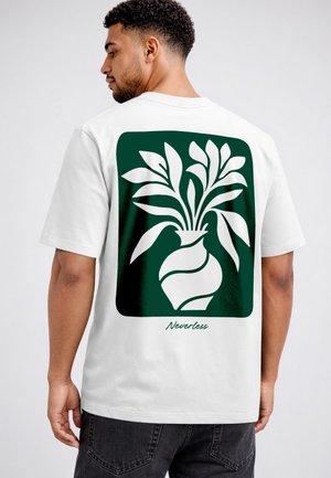 Man wearing white T-shirt with green graphic of stylized vase holding leaves and the word "Neverless" on the back.