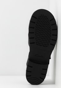 La semelle d'une chaussure noire Clarks présente un design en caoutchouc texturé avec des rainures profondes et une forme arrondie, démontrant une construction durable et une bonne adhérence.