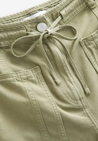 Shorts di tessuto verde chiaro con vita regolabile tramite cordino, tasche anteriori e dettagli di cucitura visibili. Texture liscia con un design informale.