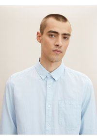 Camisa de botones de manga larga en azul claro, con cuello puntiagudo, textura sutil y un único bolsillo en el pecho con cierre de botón.