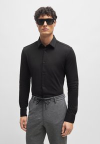 Schwarzes, langärmliges Hemd mit Knöpfen und Kragen, kombiniert mit strukturierten grauen Hosen mit Kordelzugbund. Das Model trägt schwarze Sonnenbrillen.