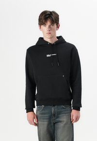 Schwarze Baumwollkapuzenjacke mit einer vorderen Tasche, Kordelzugkapuze und weißem Logotext auf der Brust. Glatte Textur und lockere Passform.