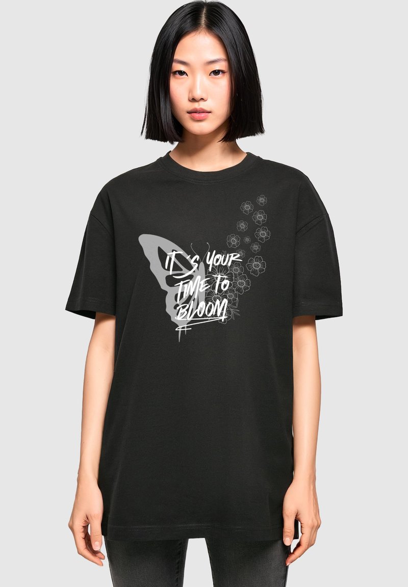 Sort bomuld t-shirt med et sommerfuglegrafik og blomsterdesigns, med hvid tekst der står "DET ER DIN TID TIL AT BLOOME" trykt på forsiden.