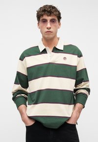 Νεαρός άνδρας που φοράει ένα ριγέ ράγκμπι πουκάμισο σε πράσινο και μπεζ χρώμα με λευκό γιακά και φιμέ γυαλιά ηλίου, τα χέρια του στις τσέπες μαύρων παντελονιών.