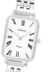 Montre rectangulaire argentée avec des index horaires en chiffres romains, cadran blanc et bracelet en métal poli. Nom de la marque "OOZOO" affiché en haut.