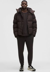 Puffer-takki tummanruskeana, jossa on huppu ja tikattu muotoilu. Yhdistetty tummiin jogging-housuihin ja musta-valkoisiin lenkkikenkiin.