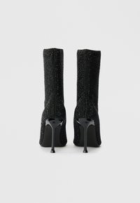 Μαύρες μπότες μέχρι το γόνατο με υφή, κρυστάλλινη φινέτσα, μυτερές μύτες και γυαλιστερές ψηλές γόβες. Ο σχεδιασμός διαθέτει σιλουέτα που αγκαλιάζει το σώμα.