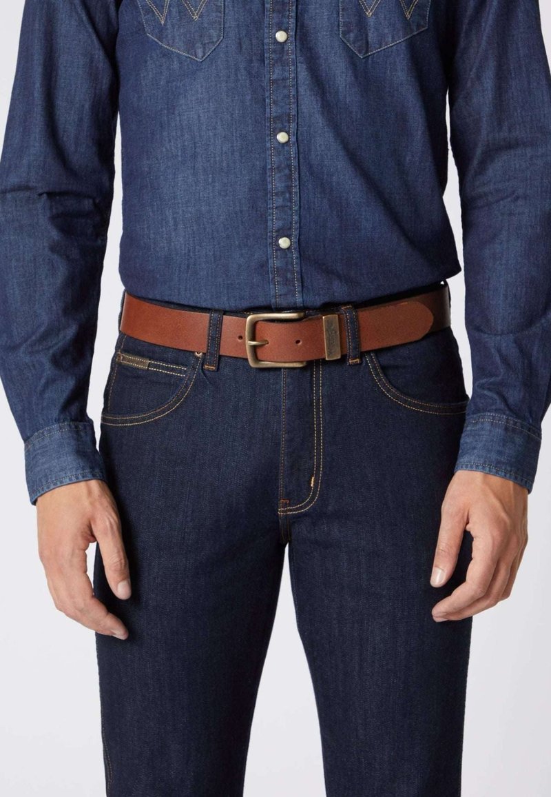 Brauner Ledergürtel mit einer metallen Schnalle, getragen über dunkelblauen Jeans und einem dunkelblauen Jeanshemd mit sichtbaren Nähten und Druckknöpfen.