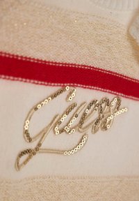 Tissu couleur crème avec une bande rouge horizontale et un logo en sequins dorés "Guess". La texture est douce avec un éclat subtil.