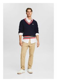Maglione navy a trecce con scollo a V e strisce rosso-bianco, indossato sopra una camicia bianca con bottoni. Chinos khaki e sneakers bianche completano il look.