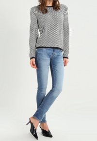 Femme portant un pull à motif noir et blanc, un jean skinny bleu délavé, et des escarpins noirs vernis à bout pointu, debout sur un fond blanc.