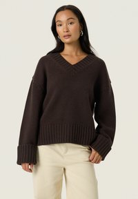 Pull en tricot brun avec un col en V, des poignets et un ourlet côtelés ; tissu texturé ; porté sur un pantalon clair.