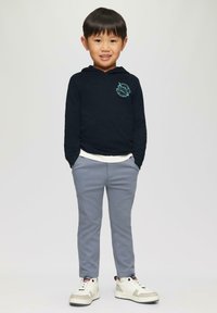 Marine hoodie met turquoise logo, gelaagd over een wit shirt; grijze broek; witte sneakers met grijze accenten; kind staat met handen in de zakken.