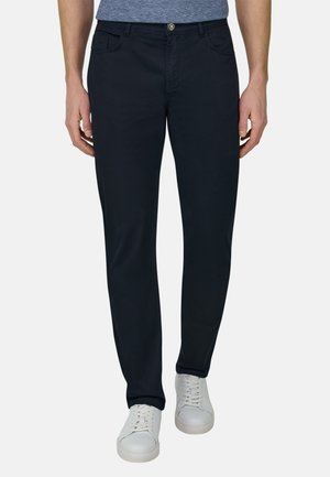 Mężczyzna ubrany w wąskie czarne spodnie, niebieską melanżową koszulę i białe sportowe trampki na sznurówki, stojący na tle gładkiego tła.