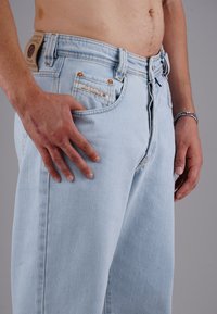 Mann in hellblauen Jeans mit aufgezipptem Reißverschluss, linke Hand auf der Hüfte, rechte Hand entspannt, zeigt den Oberkörper und den unteren Torso.