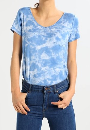 Kvinde iført en blå- og hvid tie-dye kortærmet skjorte med brystlomme og blå højtaljede jeans med hænder i lommerne.