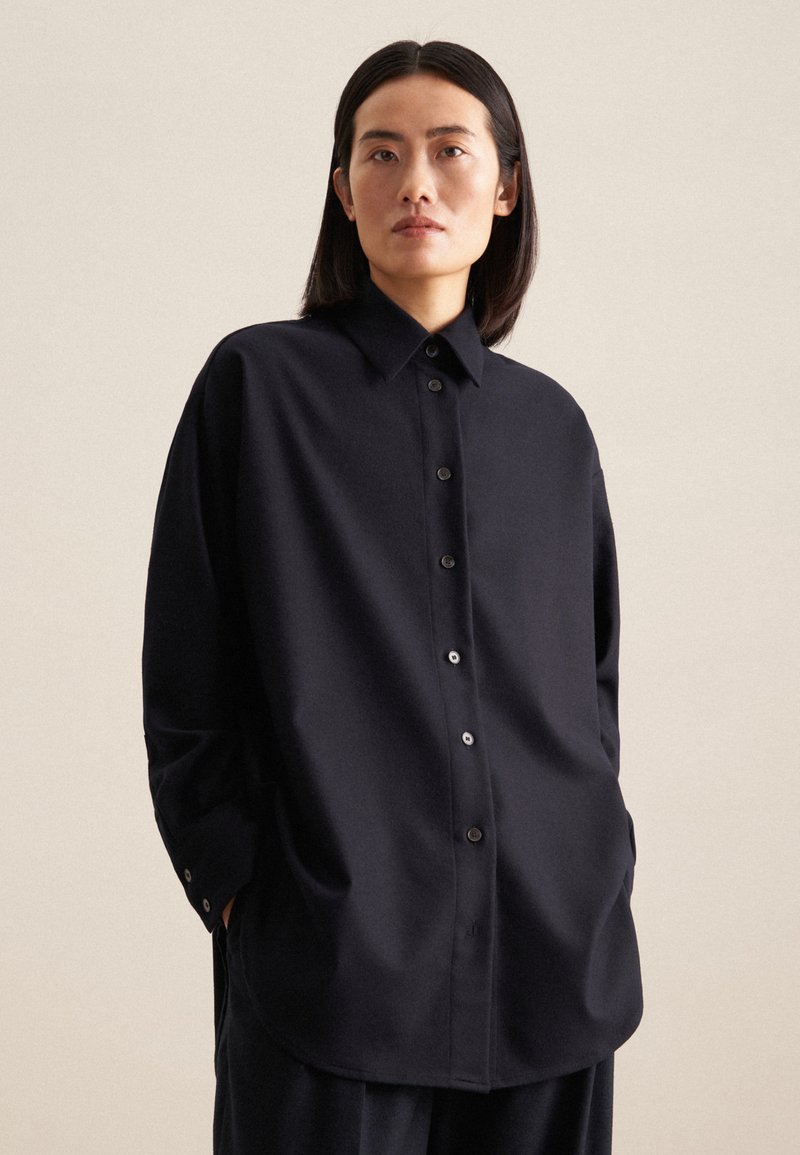 Camicia nera oversized con bottoni, realizzata in un tessuto morbido. Presenta un colletto, maniche lunghe e un orlo arrotondato con dettagli in evidenza sui bottoni.