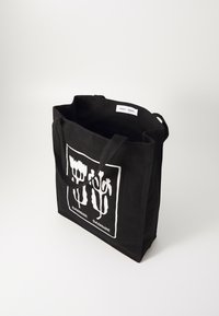Zwarte canvas tote bag met een wit bloemenontwerp, voorzien van twee stevige handvatten en een open bovenkant. Merklabel zichtbaar aan de binnenkant.