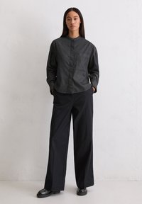 Camicia a maniche lunghe grigia scura con bottoni e tasca, abbinata a pantaloni neri a gamba larga e scarpe nere spesse, posizionata su uno sfondo neutro.