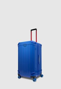 Blauer Hartschalenkoffer mit gerippter Struktur, ausziehbarem schwarzen und roten Griff, vier blauen Rollen und seitlichem Griff.