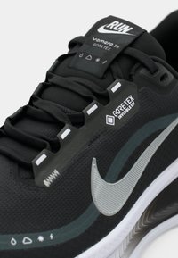 Nike Vomero 18 sportcipők fekete és szürke színben, texturált felsőrésszel, Gore-Tex címkével, fényvisszaverő logóval és vastag fehér talppal.