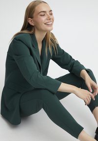 Mörkgrön skräddarsydd blazer och matchande avsmalnande byxor, tillverkade av ett glatt tyg. Har en enda knappstängning och slim fit.
