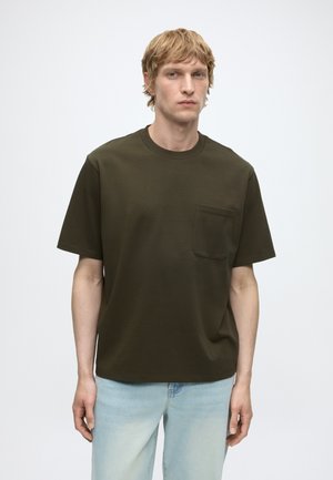 Uomo biondo che indossa una maglietta a maniche corte verde oliva con taschino sul petto e jeans azzurri, in piedi davanti a uno sfondo semplice.