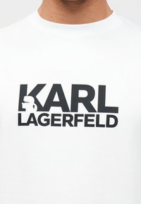 Λευκό T-shirt με έντονο μαύρο κείμενο "KARL LAGERFELD" και σιλουέτα προφίλ ενός προσώπου με γυαλιά ηλίου και κοτσίδα μέσα στο γράμμα "K".