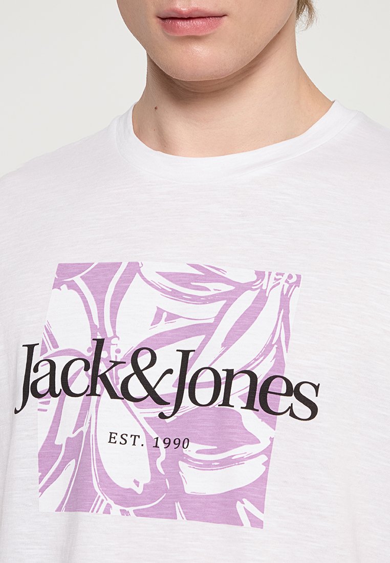 Camiseta blanca de manga corta con un gráfico floral morado y un texto en negrita negro "Jack & Jones EST. 1990" en el centro. Material de algodón suave.