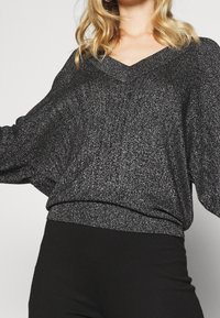 Czarny, metaliczny sweter z dzianiny z dekoltem w kształcie V, rękawami nietoperzowymi i dopasowanym ściągaczem; ma błyszczącą teksturę.